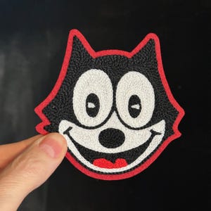 Op de afbeelding: Geborduurde patch met Felix de Kat. De patch toont een zwarte kattenkop met witte ogen, een witte mond en een rode rand. De patch wordt tegen een zwarte achtergrond gehouden.