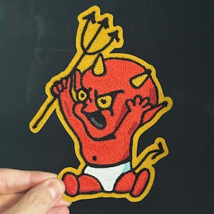 May include: An embroidered patch featuring a red devil character with yellow horns and eyes, wearing a white diaper. The character holds a black trident and is outlined in yellow thread, creating a bold, colorful design for fabric application.