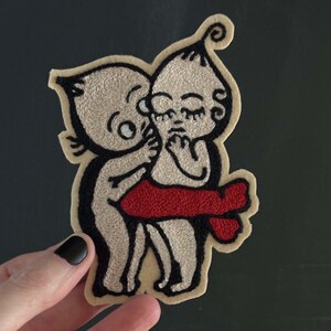 May include: Embroidered felt patch featuring two figures, one in red and one in beige, embracing. The figures are stylized and have simple features. The patch is perfect for adding a unique touch to clothing, bags, or other accessories.