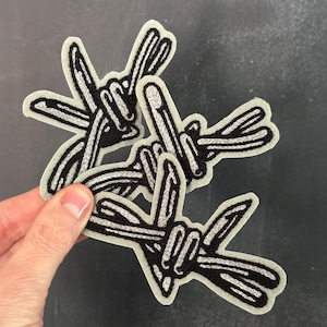 Może przedstawiać: Trzy haftowane naszywki z czarno-białym wzorem drutu kolczastego. Naszywki mają jasnoszarą obwódkę i są trzymane w dłoni na ciemnym tle. Te naszywki to wyjątkowy dodatek.