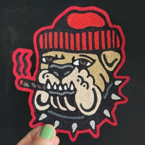 Könnte beinhalten: Ein bestickter Aufnäher mit einem Bulldoggen-Motiv, die eine rot-schwarz gestreifte Mütze trägt. Die Bulldogge hat ein beiges Gesicht, ein schwarzes Halsband mit silbernen Nieten und hält eine brennende Zigarre mit rotem Rauch im Maul.