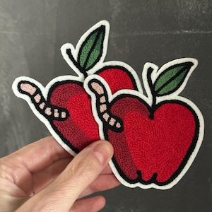 Op de afbeelding: Geborduurde appelpatches met een wormontwerp. De appels zijn rood met een zwarte omtrek en een groen blad. De worm is roze en wit. De patches hebben een witte rand.