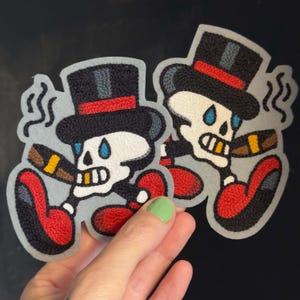 May include: Two embroidered patches featuring a cartoon skull wearing a black top hat with a red band. The skull has blue teardrops under the eyes, a cigar in its mouth, and wears large red shoes. The design includes grey smoke swirls.