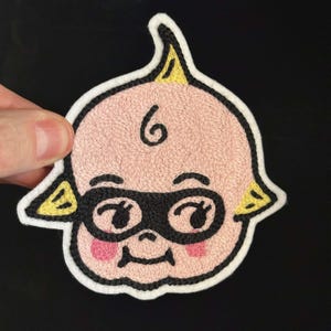 May include: Embroidered patch featuring a baby face design. The patch has a pink face with black eye mask, eyebrows, and eyes. The baby has a swirl of hair and yellow accents. The patch is outlined in white.