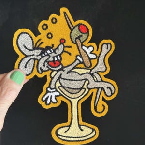 Puede incluir: Un parche bordado que muestra un ratón de dibujos animados gris descansando dentro de una copa de martini de color amarillo claro. El ratón tiene la boca roja y sostiene un palillo con una aceituna verde. El diseño tiene un borde amarillo mostaza.