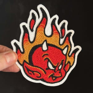 May include: Embroidered patch featuring a red devil with white horns and a mischievous expression, surrounded by orange and yellow flames. The patch has a white border and black outline, perfect for jackets, bags, or other fabric items.