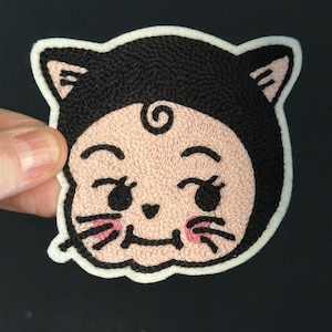Op de afbeelding: Geborduurde patch met een cartoonachtig katachtig karakter met een zwarte capuchon en een knipogende uitdrukking. Het gezicht is lichtroze met zwarte ogen, snorharen en een kleine mond. De patch heeft een witte rand.