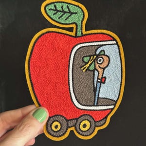 Op de afbeelding: Een rode, appelvormige geborduurde patch met een groen blaadje aan de bovenkant en twee zwarte wielen aan de onderkant. Binnenin de appel onthult een lichtblauw venster een beige worm met een groene hoed en een blauw overhemd met rode vlinderdas.