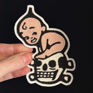 Puede incluir: Parche bordado con una figura de bebé sobre una calavera. El bebé está representado en hilo rosa y negro, con una expresión sonriente. La calavera es blanca con contornos negros, y el parche tiene un borde blanco. Para ropa o accesorios.