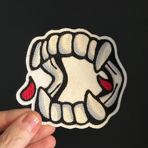 May include: Embroidered patch featuring a stylized set of vampire fangs. The design includes white teeth with black outlines, a white background, and two red blood drops. The patch is likely for clothing or accessories.