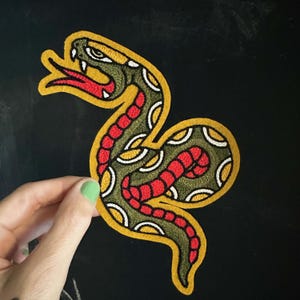 May include: An embroidered patch shaped like a coiled snake with a yellow border. The snake features a dark olive green body with red scales, white accents, and a bright red forked tongue, all rendered in a detailed, textured thread design.