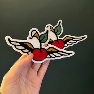 Puede incluir: Dos parches bordados de cerezas rojas con tallos y hojas verdes, cada uno con alas blancas. Las alas están delineadas en negro y tienen un acento amarillo.