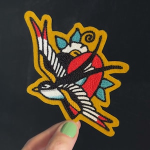 May include: An embroidered patch featuring a stylized swallow in flight with black, white, and red plumage. The bird is positioned against a circular red background with teal leaves and a yellow border, creating a vibrant, multi-colored design.