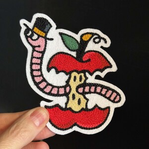Op de afbeelding: Geborduurde patch met een worm met hoge hoed die uit een rode appel komt. De appel heeft een hap genomen, waardoor een gele kern zichtbaar wordt. De worm is roze en de appel heeft een groen blad.