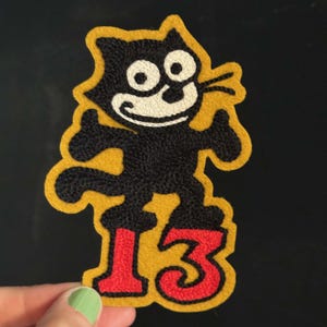 May include: An embroidered patch featuring a black cartoon cat with a white face and wide eyes, standing on top of the red number 13. The entire design is outlined in a mustard yellow border, creating a distinct shape against the dark background.
