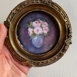 May include: A framed painting of a bouquet of pink flowers in a blue vase. The flowers are painted in a realistic style and the background is a dark purple.