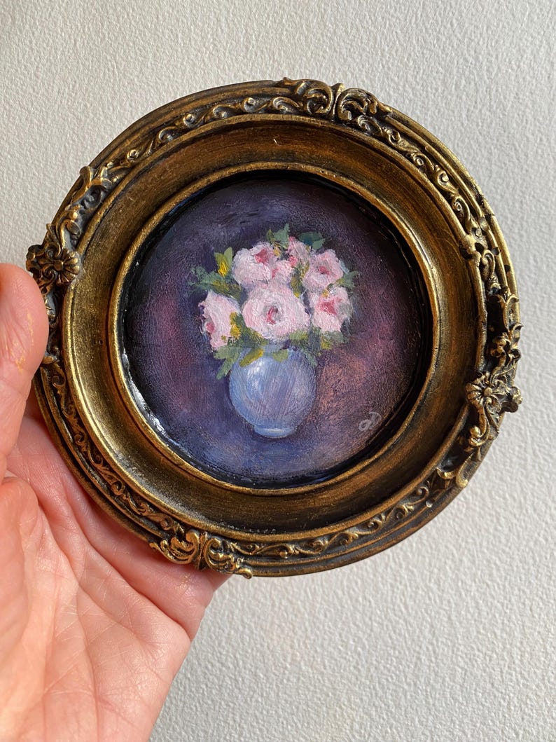 May include: A framed painting of a bouquet of pink flowers in a blue vase. The flowers are painted in a realistic style and the background is a dark purple.