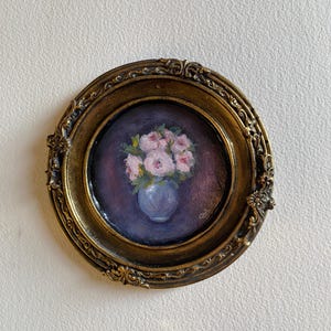 May include: A framed painting of a bouquet of pink roses in a blue vase. The painting is set in a gold oval frame with ornate details.