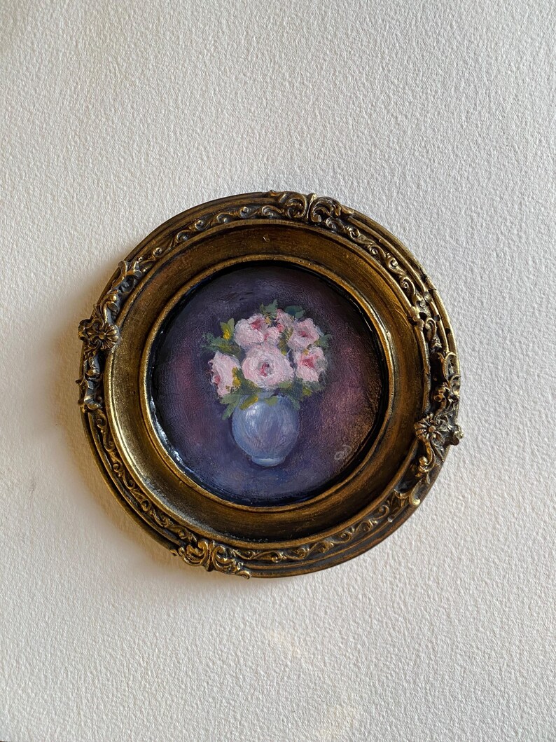 May include: A framed painting of a bouquet of pink roses in a blue vase. The painting is set in a gold oval frame with ornate details.