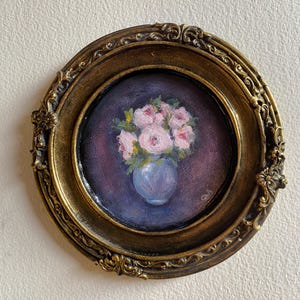 May include: A framed painting of a bouquet of pink flowers in a blue vase. The painting is set against a dark purple background.