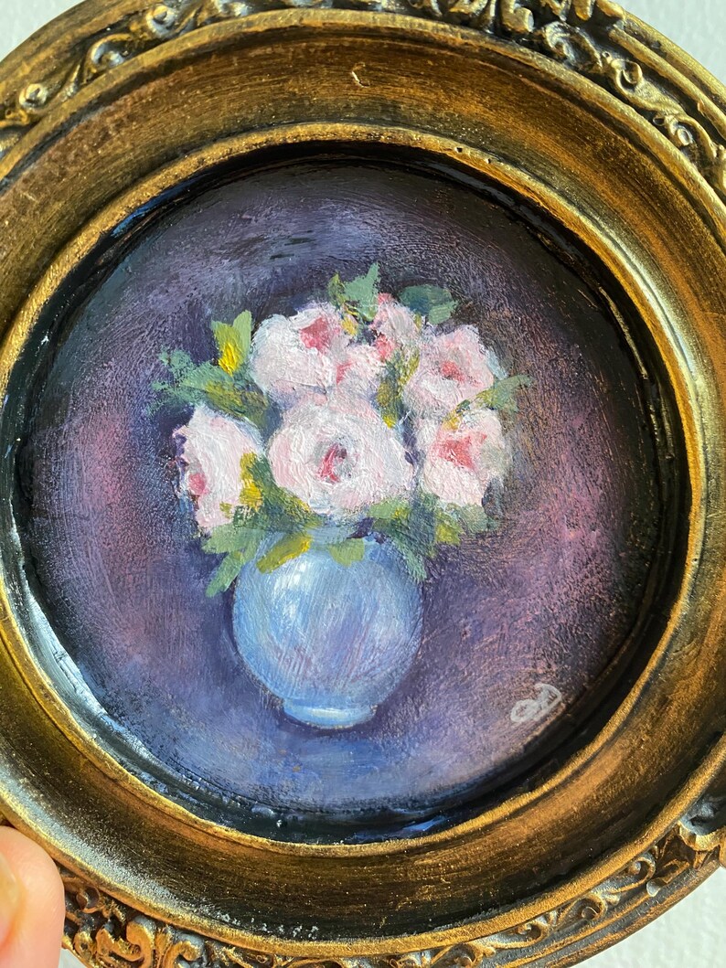 May include: A framed painting of a bouquet of pink flowers in a blue vase. The flowers are painted in a realistic style and the background is a dark purple.