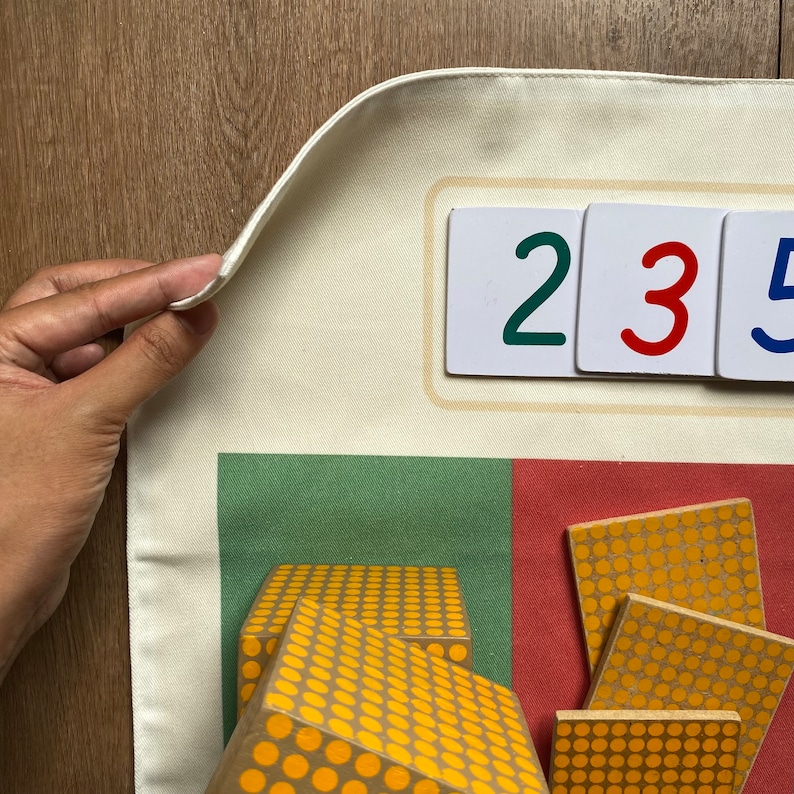 May include: A white canvas play mat with a green and red section. The mat has three white cards with the numbers 2, 3, and 5 printed in different colors. There are also wooden blocks with yellow dots on them.