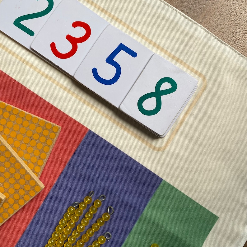 May include: A white fabric mat with colorful number cards on top. The numbers are 2, 3, 5, and 8. The numbers are written in different colors. There are yellow beads on a blue surface below the cards.