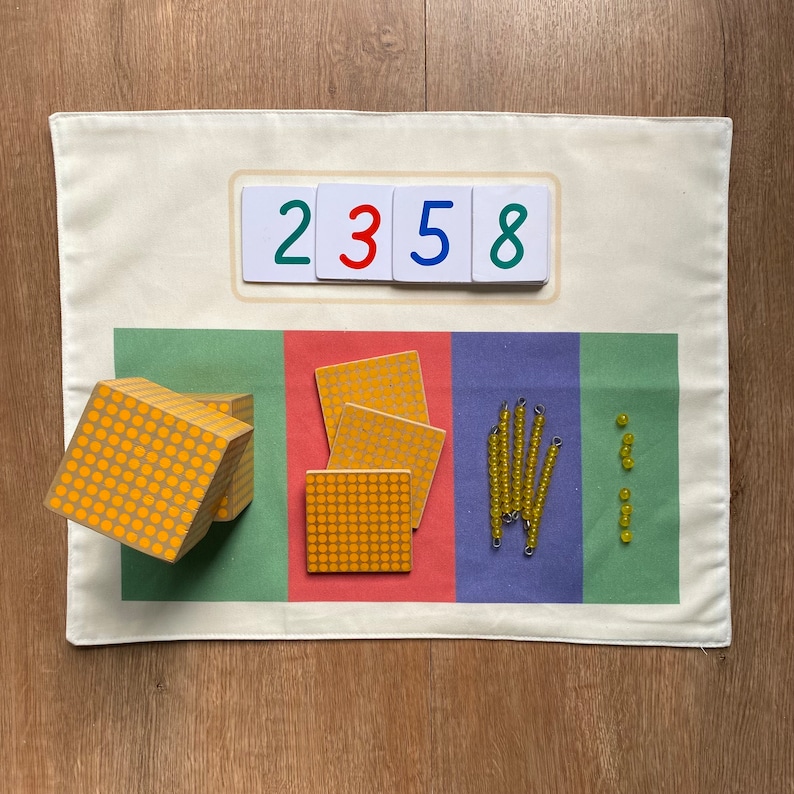 May include: A white fabric mat with a green, red, and blue section. Each section has different Montessori learning materials. The green section has a yellow wooden cube with yellow dots. The red section has three yellow squares with yellow dots. The blue section has five yellow bead strings and a row of yellow beads. The top of the mat has white cards with the numbers 2, 3, 5, and 8.