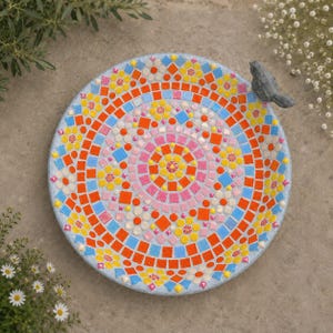 May include: A circular bird bath featuring a vibrant mosaic pattern of orange, yellow, pink, and blue tiles arranged in concentric rings. A small, dark grey bird figurine is attached to the rim of the basin, which has a light grey base.