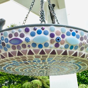 May include: A hanging bird bath featuring a mosaic design with blue, purple, and white tiles in circular and triangular patterns. A small gray bird figurine is attached to the rim. The basin is suspended by metal chains from a white structure.