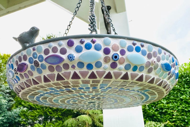 May include: A hanging bird bath featuring a mosaic design with blue, purple, and white tiles in circular and triangular patterns. A small gray bird figurine is attached to the rim. The basin is suspended by metal chains from a white structure.
