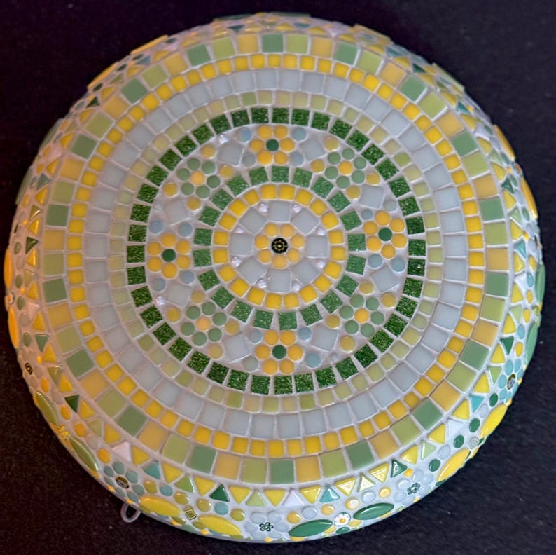 May include: A circular mosaic wall art piece featuring a radial pattern of small square and triangular tiles in shades of yellow, light green, dark green, and white. The design includes concentric rings with floral motifs and glittery accents.