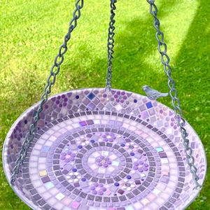 May include: A circular hanging bird bath featuring a mosaic design in various shades of purple, lavender, and white. The surface is composed of small square and floral-shaped tiles arranged in concentric patterns, with a small gray bird figurine.
