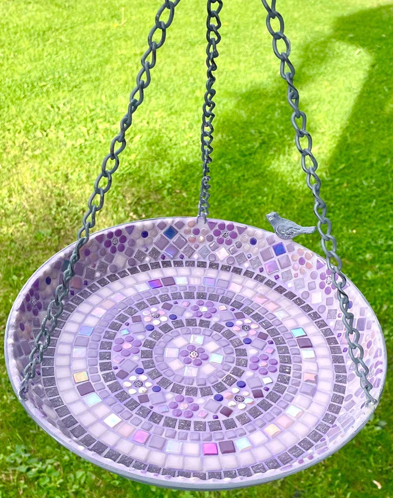 May include: A circular hanging bird bath featuring a mosaic design in various shades of purple, lavender, and white. The surface is composed of small square and floral-shaped tiles arranged in concentric patterns, with a small gray bird figurine.