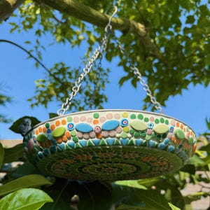 May include: A round hanging bird bath featuring a mosaic exterior made of small green, orange, pink, and blue tiles. The basin is suspended by a metal chain and includes a small decorative bird figure attached to the outer rim of the bowl.