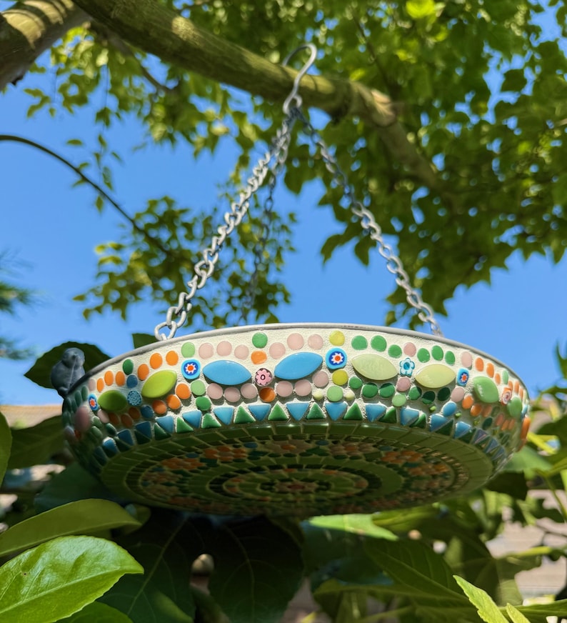 May include: A round hanging bird bath featuring a mosaic exterior made of small green, orange, pink, and blue tiles. The basin is suspended by a metal chain and includes a small decorative bird figure attached to the outer rim of the bowl.