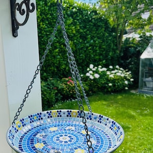 May include: A round hanging bird bath featuring a mosaic interior with blue, white, and yellow tiles arranged in a circular pattern. The basin is suspended by three metal chains attached to a black wrought iron wall bracket and includes a small black bird figurine perched on the rim.