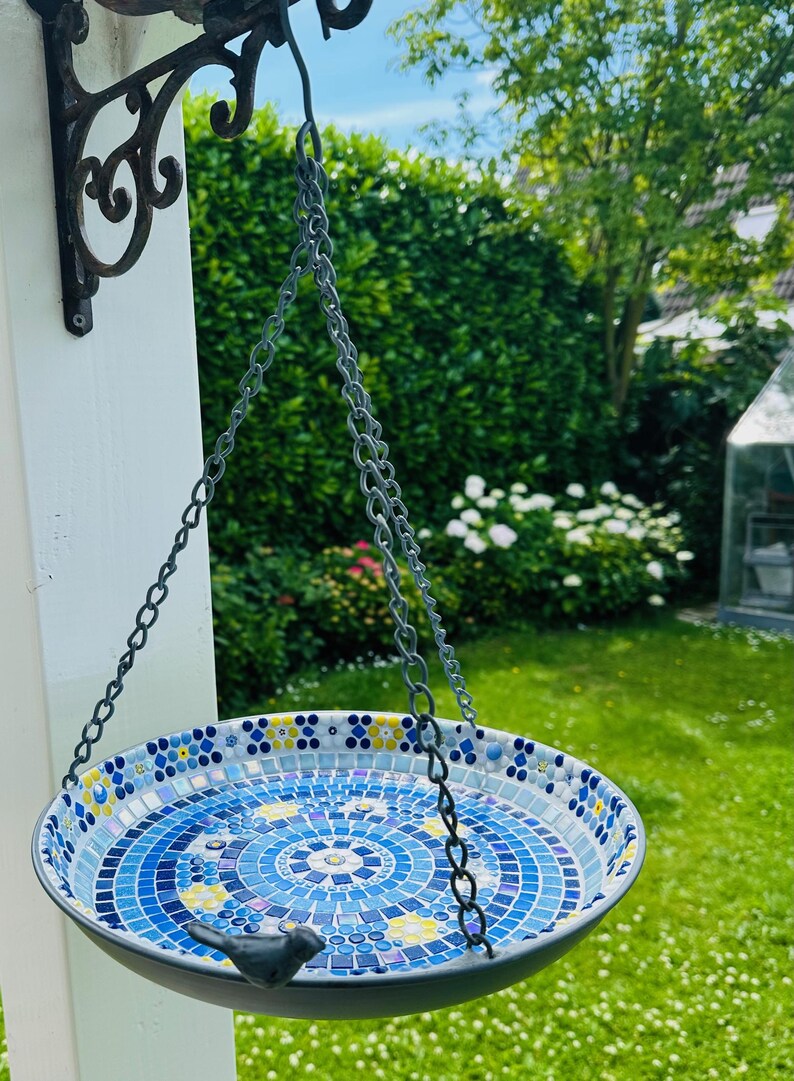 May include: A round hanging bird bath featuring a mosaic interior with blue, white, and yellow tiles arranged in a circular pattern. The basin is suspended by three metal chains attached to a black wrought iron wall bracket and includes a small black bird figurine perched on the rim.