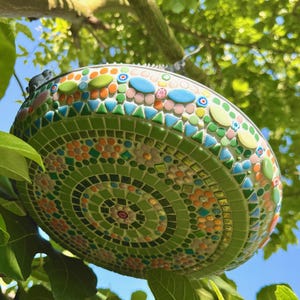 May include: A round mosaic bird bath featuring a vibrant pattern of green, orange, pink, and blue glass tiles. The surface is decorated with concentric circles and floral motifs made from small square, triangular, and oval-shaped glass pieces.