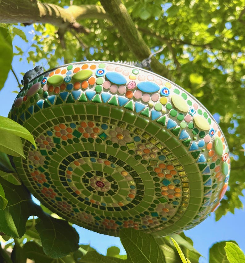May include: A round mosaic bird bath featuring a vibrant pattern of green, orange, pink, and blue glass tiles. The surface is decorated with concentric circles and floral motifs made from small square, triangular, and oval-shaped glass pieces.
