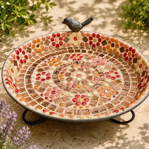 May include: A round bird bath featuring a mosaic pattern of red, orange, and cream-colored tiles arranged in floral and geometric designs. A small, dark gray metal bird figurine is attached to the rim, and the basin rests on a black metal stand.