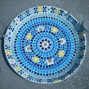 May include: A round bird bath featuring a mosaic design of blue, light blue, and yellow tiles arranged in concentric circles. The pattern includes small floral motifs and a small gray bird figurine attached to the outer rim of the basin.