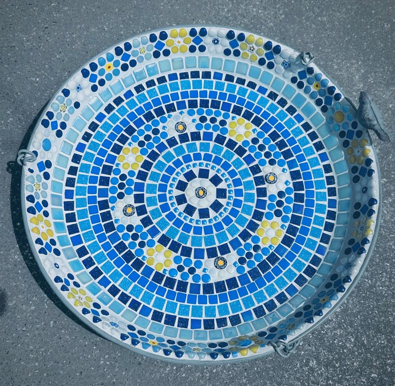 May include: A round bird bath featuring a mosaic design of blue, light blue, and yellow tiles arranged in concentric circles. The pattern includes small floral motifs and a small gray bird figurine attached to the outer rim of the basin.