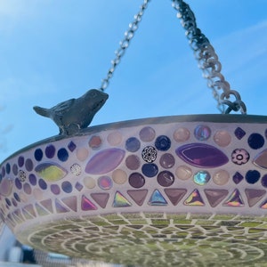 May include: A hanging bird bath featuring a mosaic design with purple, pink, and iridescent glass tiles. A small dark gray bird figurine is attached to the rim. The basin is suspended by a metal chain against a clear blue sky background.