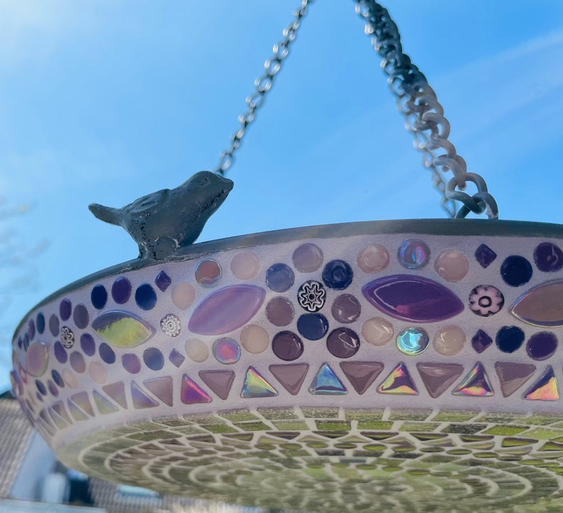 May include: A hanging bird bath featuring a mosaic design with purple, pink, and iridescent glass tiles. A small dark gray bird figurine is attached to the rim. The basin is suspended by a metal chain against a clear blue sky background.