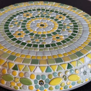 May include: A circular mosaic trivet featuring a radial pattern of small glass tiles in shades of yellow, lime green, forest green, and white. The design incorporates floral motifs and geometric shapes arranged in concentric rings with grout.