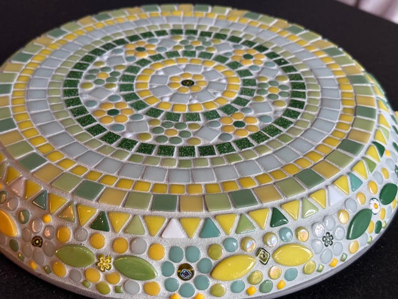 May include: A circular mosaic trivet featuring a radial pattern of small glass tiles in shades of yellow, lime green, forest green, and white. The design incorporates floral motifs and geometric shapes arranged in concentric rings with grout.