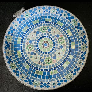 May include: A circular mosaic birdbath featuring a geometric pattern of blue, light blue, and pale green glass tiles. The design includes small floral motifs and a small, light-colored bird figurine attached to the upper left rim of the basin.