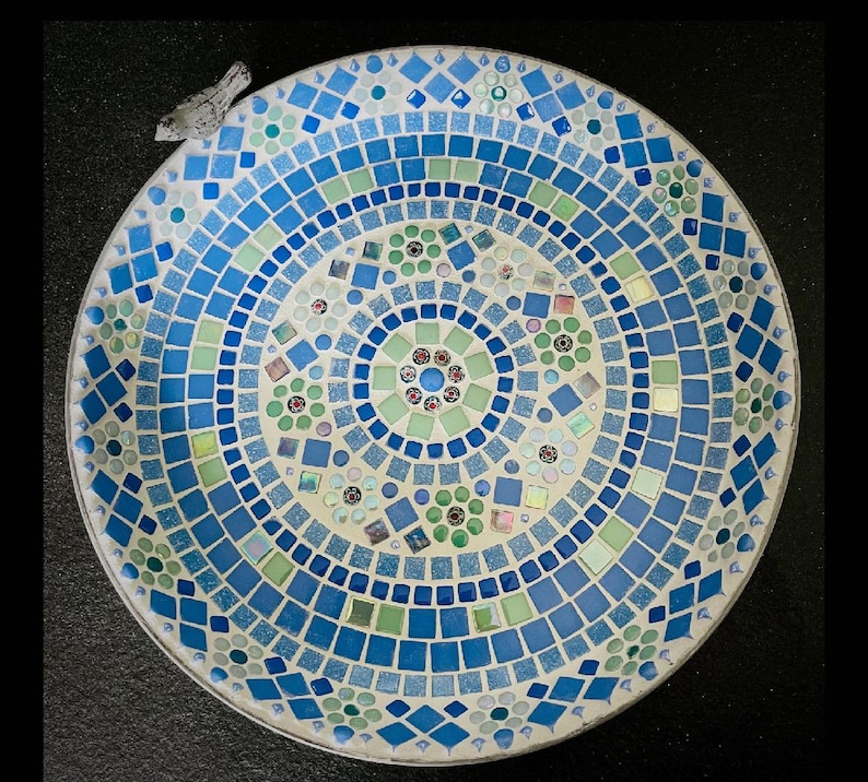 May include: A circular mosaic birdbath featuring a geometric pattern of blue, light blue, and pale green glass tiles. The design includes small floral motifs and a small, light-colored bird figurine attached to the upper left rim of the basin.