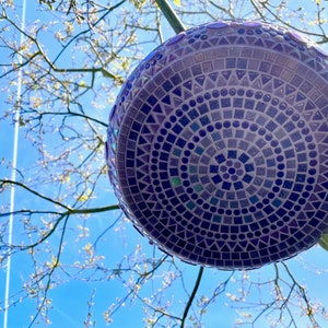 May include: A spherical garden ornament featuring a detailed mosaic pattern composed of small purple, blue, and dark violet tiles in circular, square, and triangular shapes, creating a geometric design across the entire surface of the object.